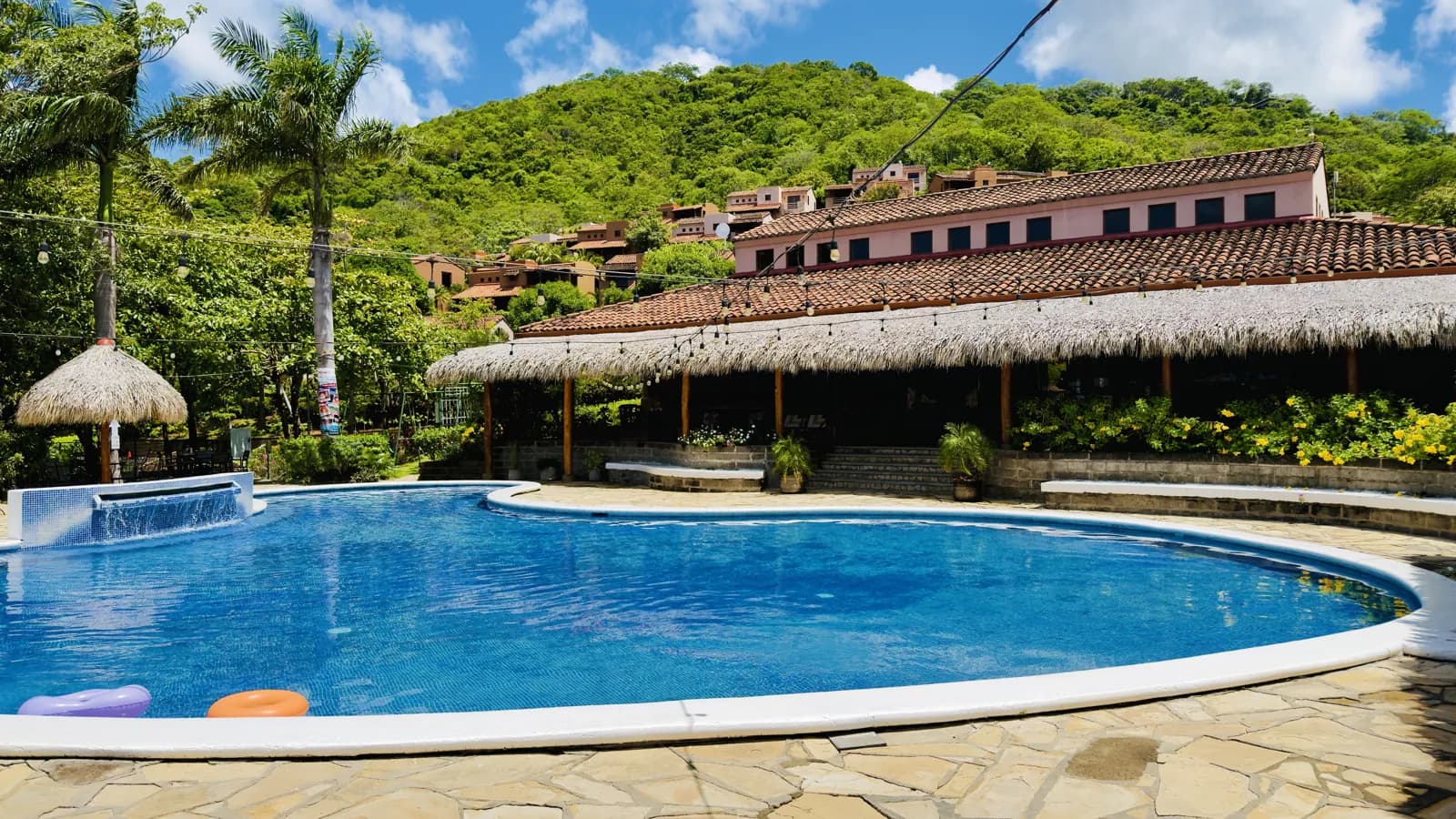 Terraza de Palermo bajo su techo de palapa junto a la piscina, con las villas en la colina